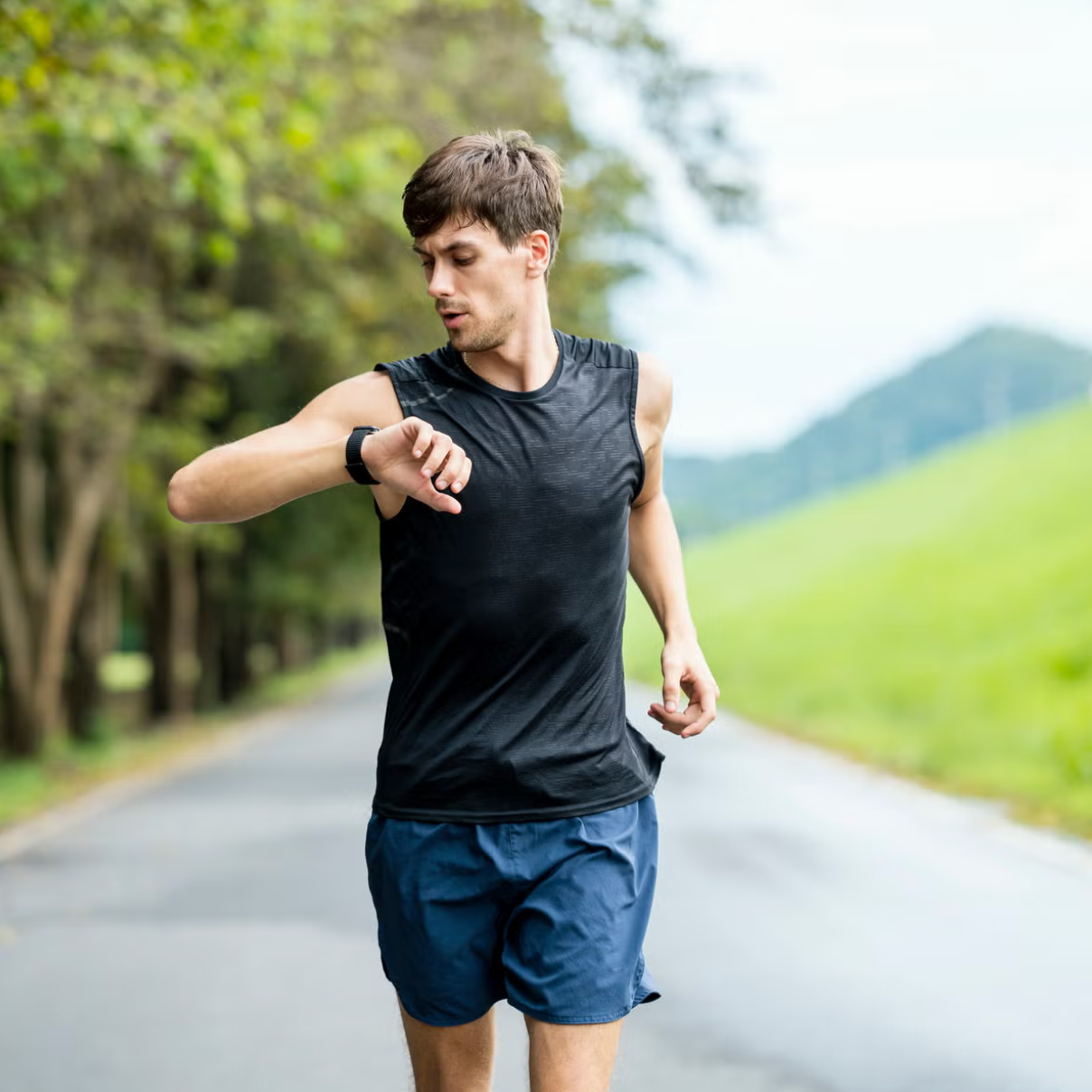 Runner checking his watch during a run on a tree-lined road with green hills in the background