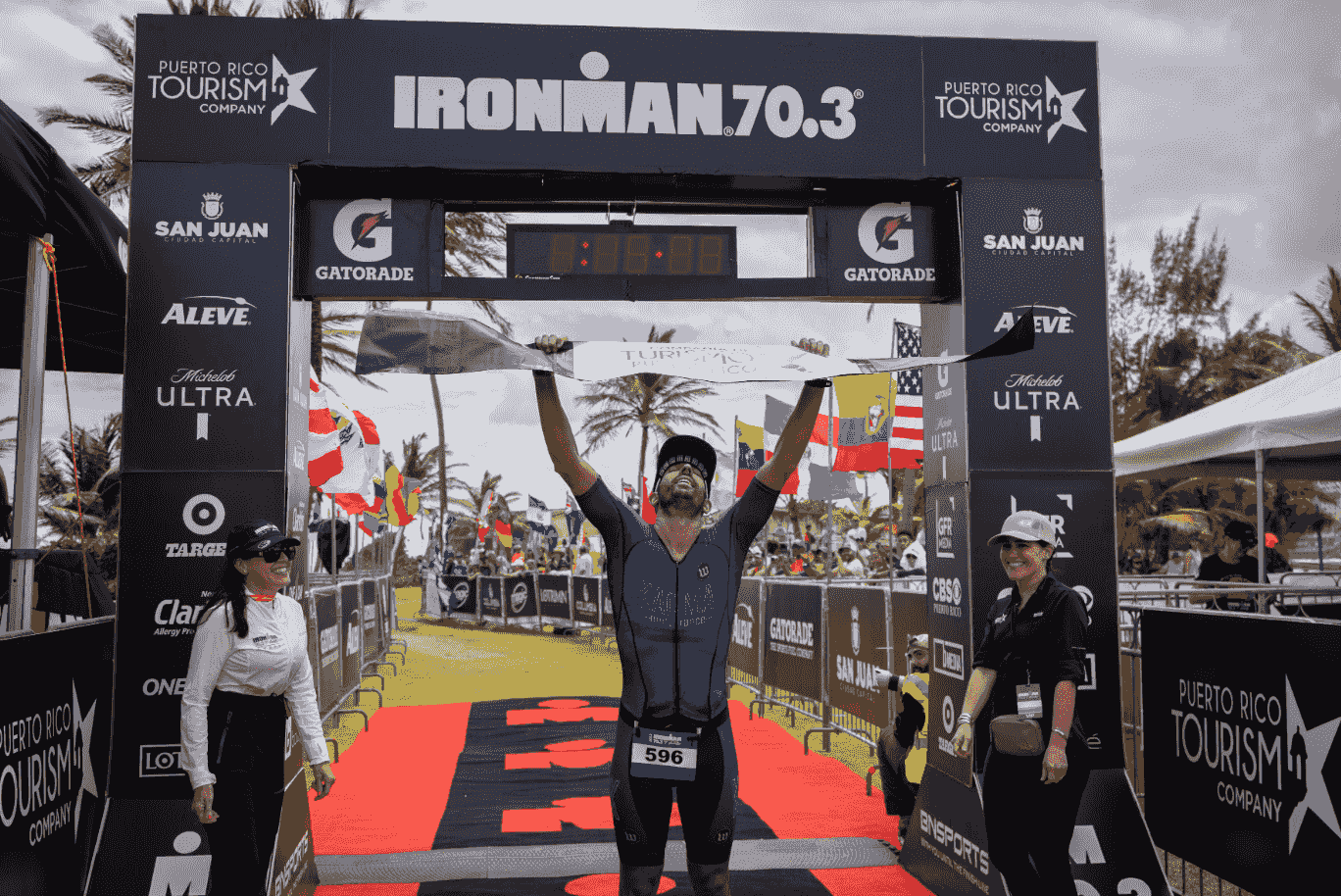 Breno Melo crossing the finish line as the winner of the 2022 Ironman 70.3 Puerto Rico, holding the banner under the clock