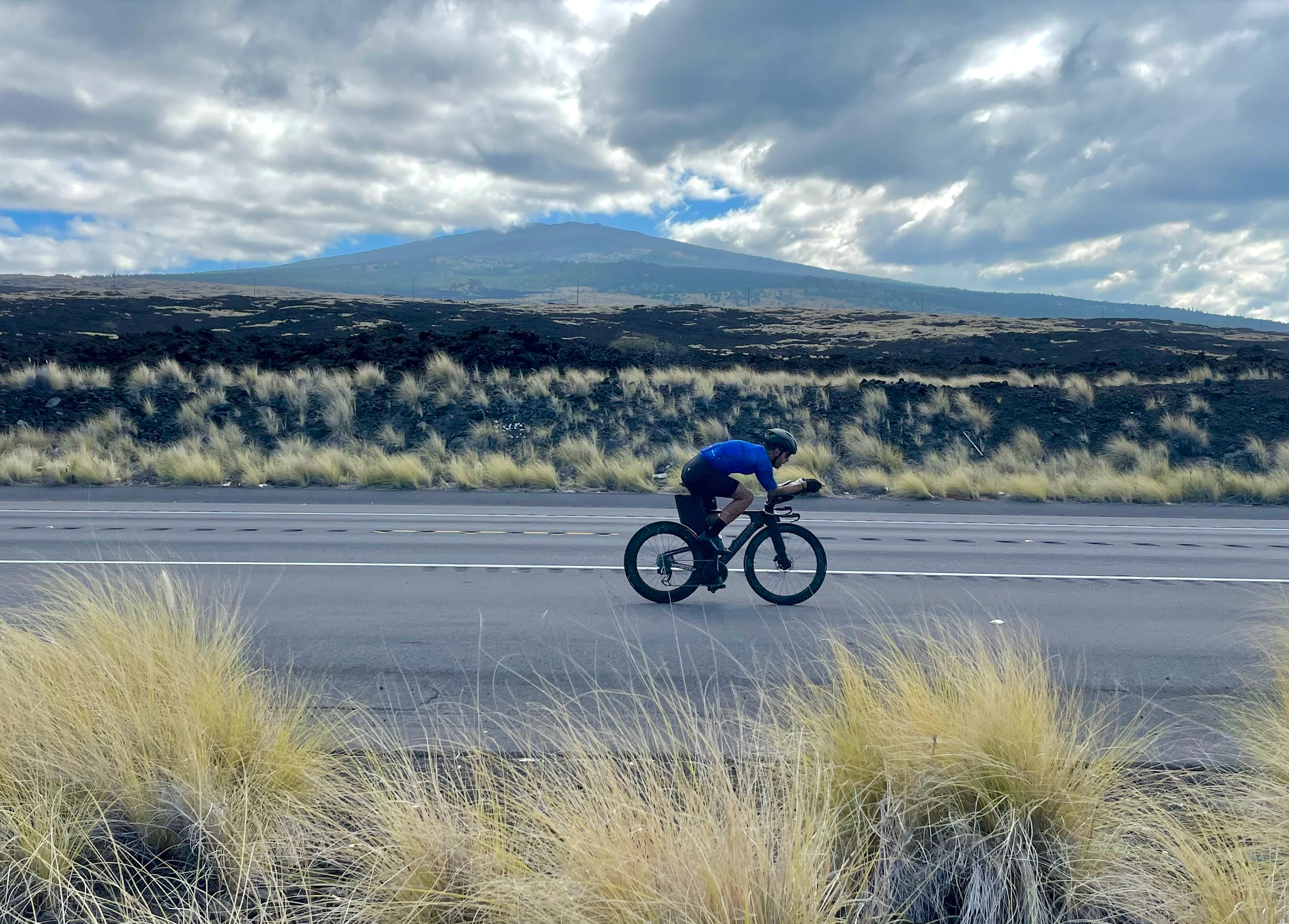 Triathlete riding a bicycle on a scenic highway in Hawaii with volcanic landscape and mountains in the background under a cloudy sky