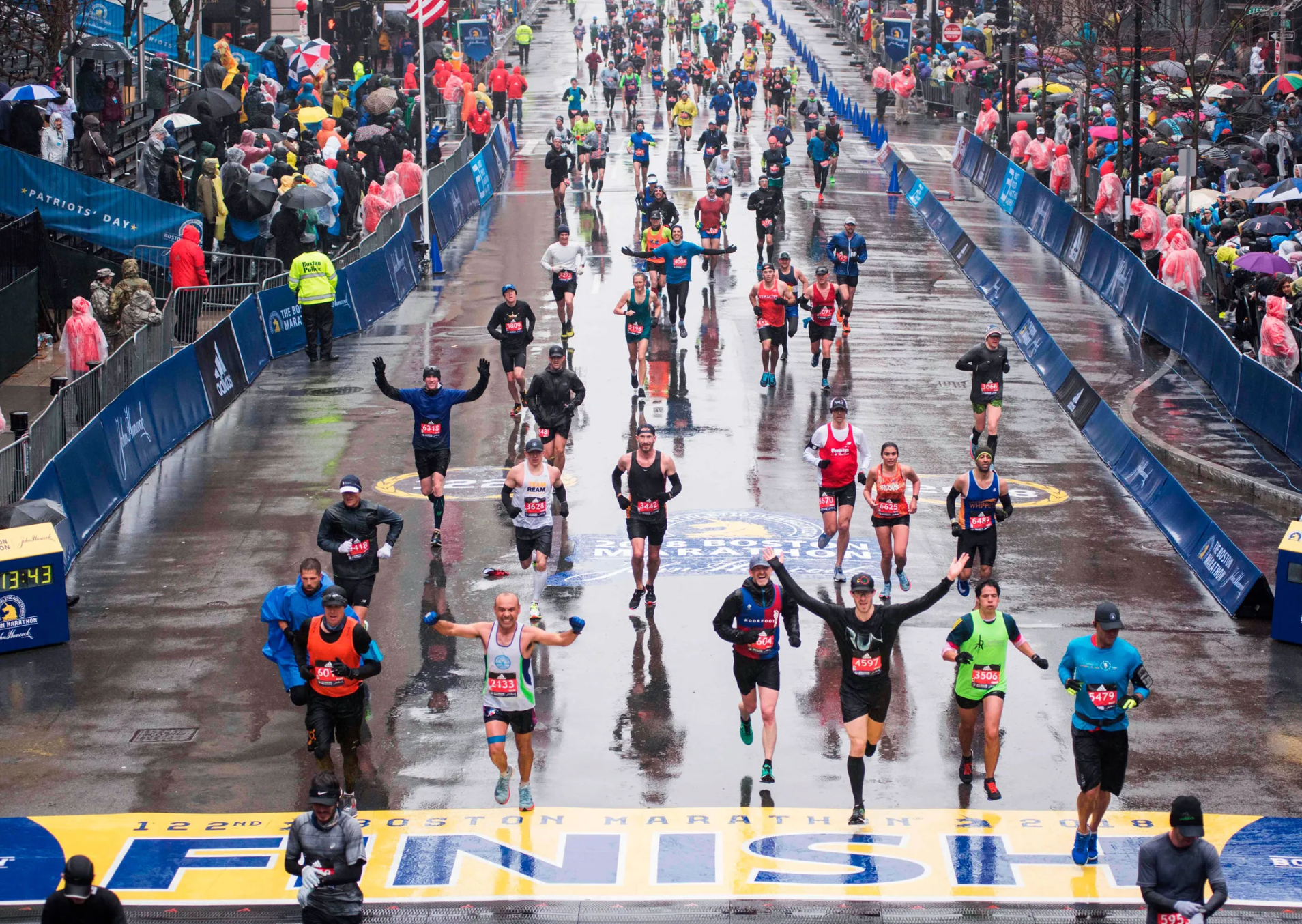 Runners crossing the finish line of the Boston Marathon on a rainy day, with cheering spectators along the sidelines