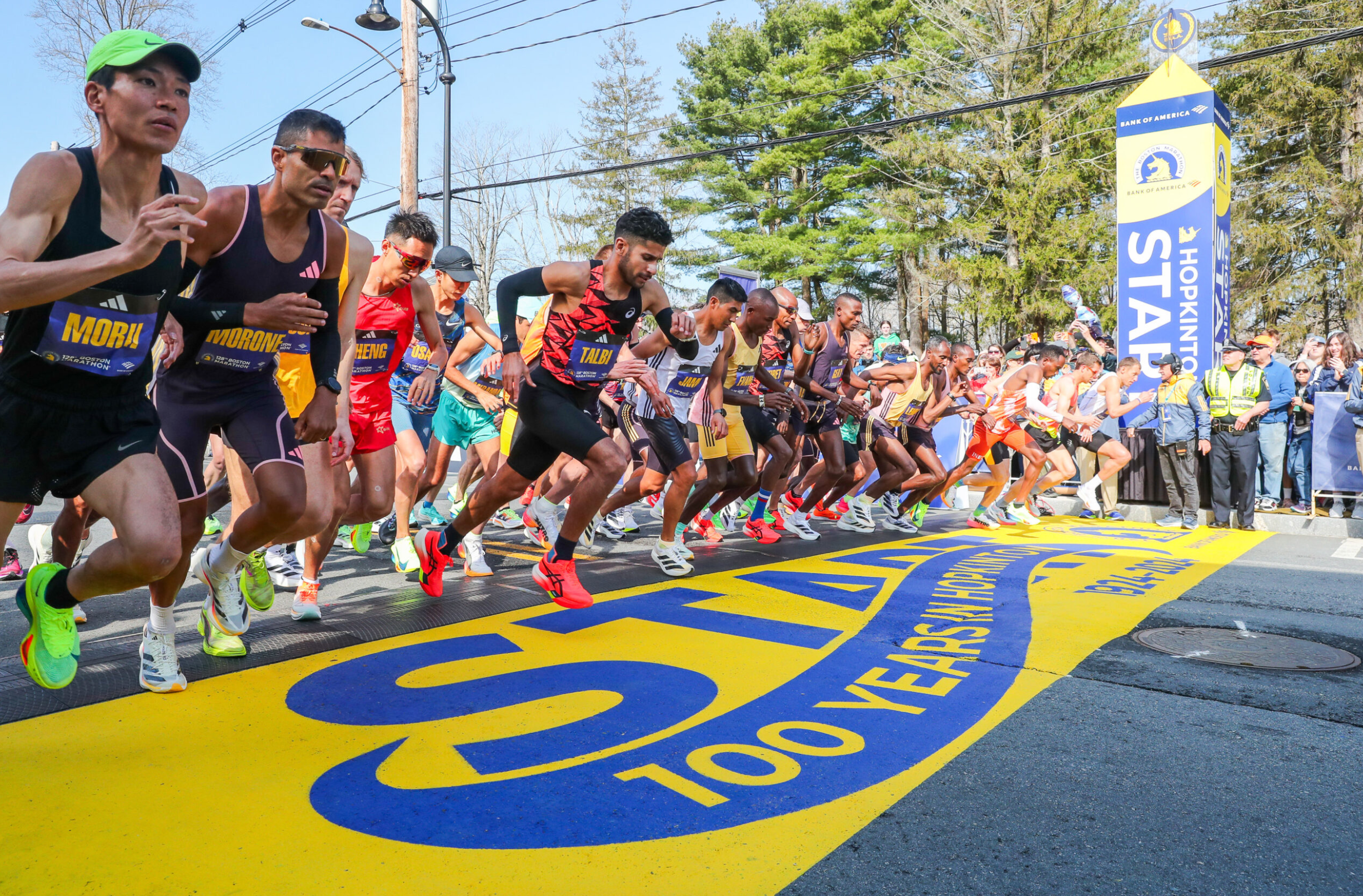 Elite marathon runners starting the race at the Boston Marathon with the Hopkinton Start line in view and '100 Years' celebration marking on the ground.