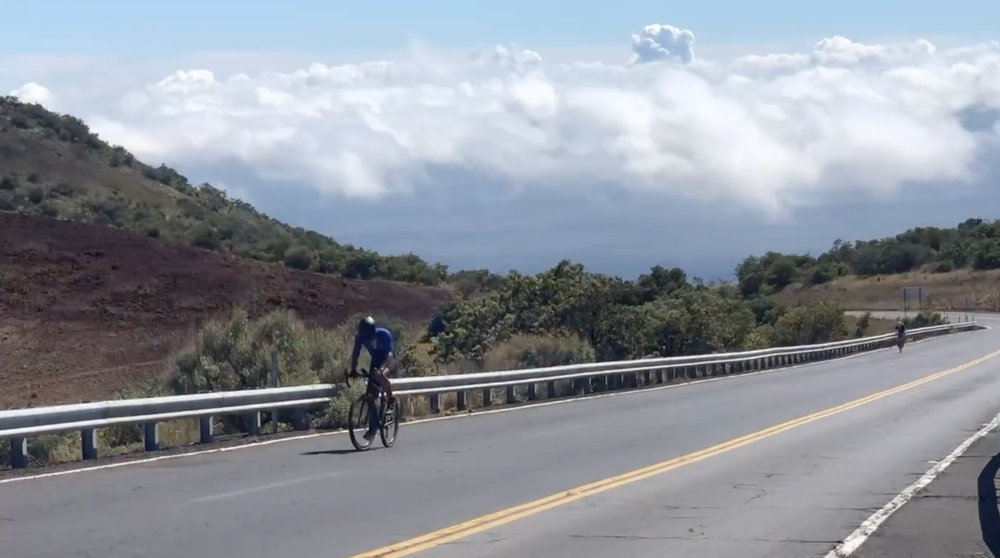 Cyclist riding on a mountain road in Hawaii with scenic views of hills, greenery, and clouds in the background, capturing a steep climb