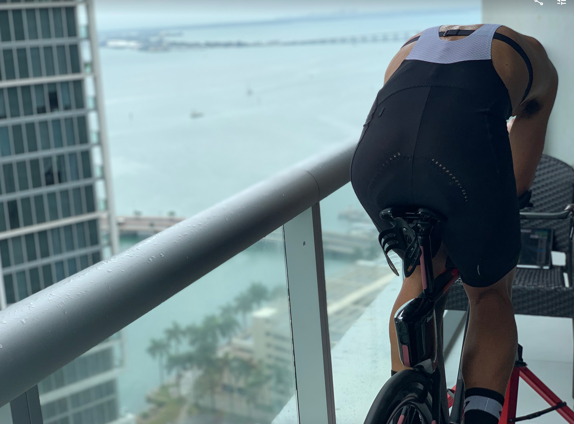 Athlete cycling on a trainer on a high-rise balcony in Miami overlooking a cityscape and waterfront, with a view of buildings, palm trees, and the ocean in the background