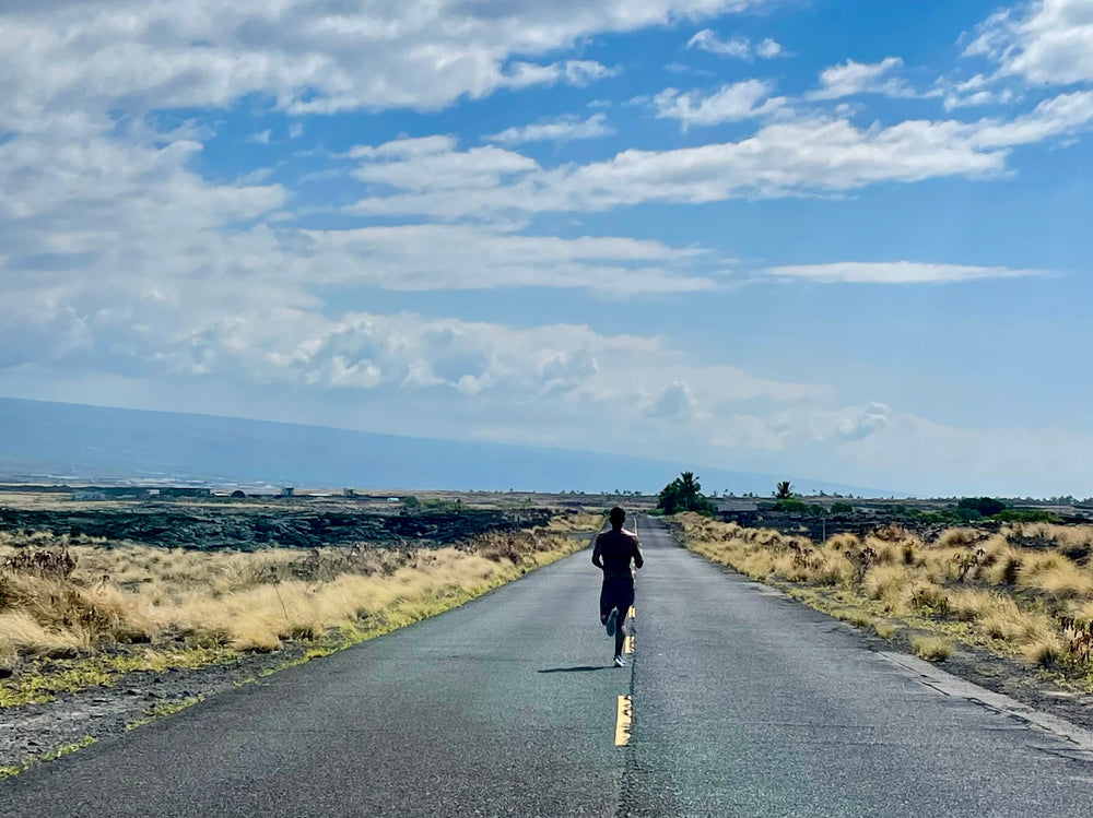 Runner on a quiet road in Hawaii, surrounded by volcanic landscape and grasslands, with a view of mountains and a partly cloudy sky