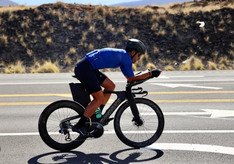 Cyclist in a blue jersey riding a time-trial bike beside dark rocky terrain